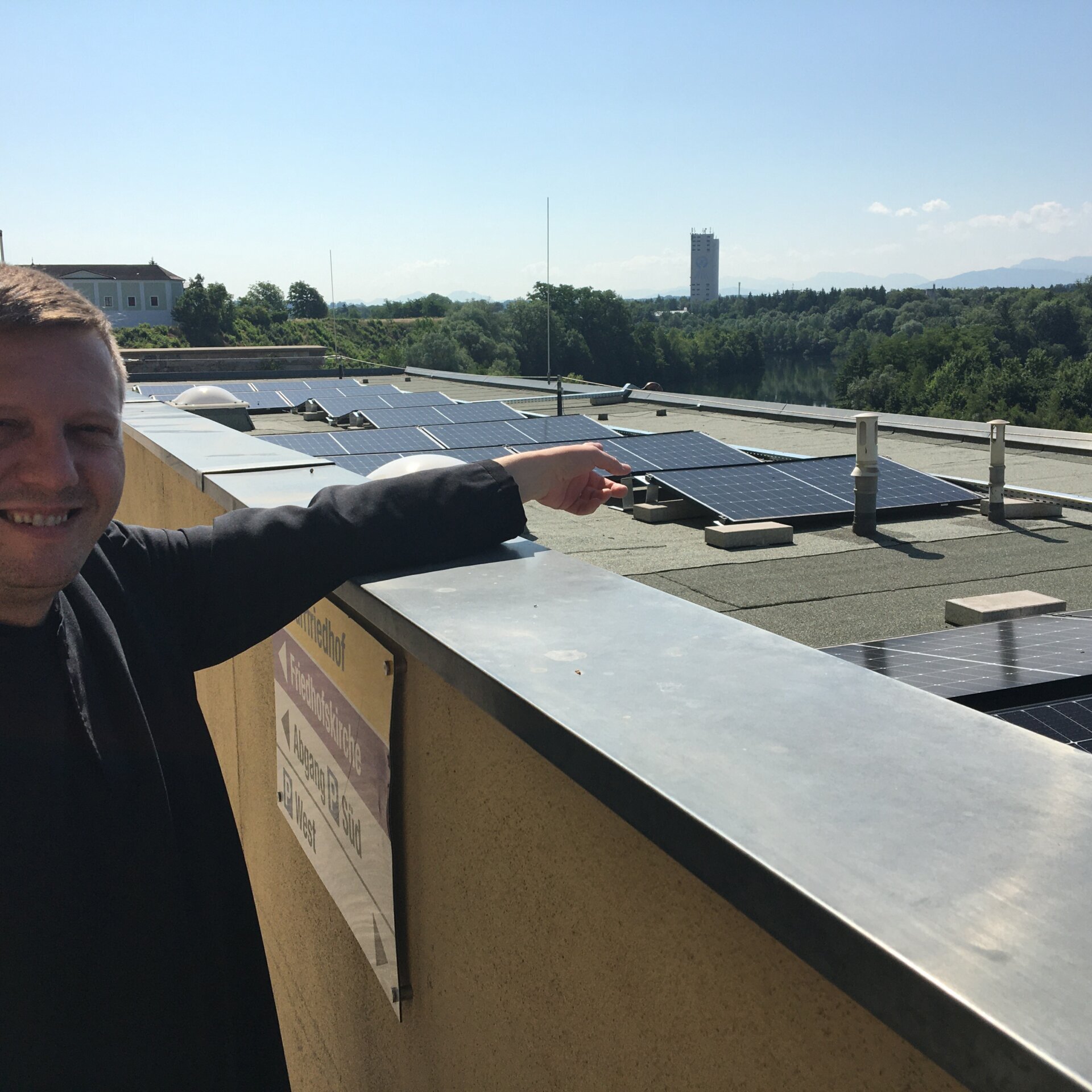 26-06-2023PV Anlage (10) Ein Mann steht auf einem Dach, lächelt und zeigt auf die auf dem Dach installierten Solarzellen, während im Hintergrund Bäume und ein Gebäude zu sehen sind.