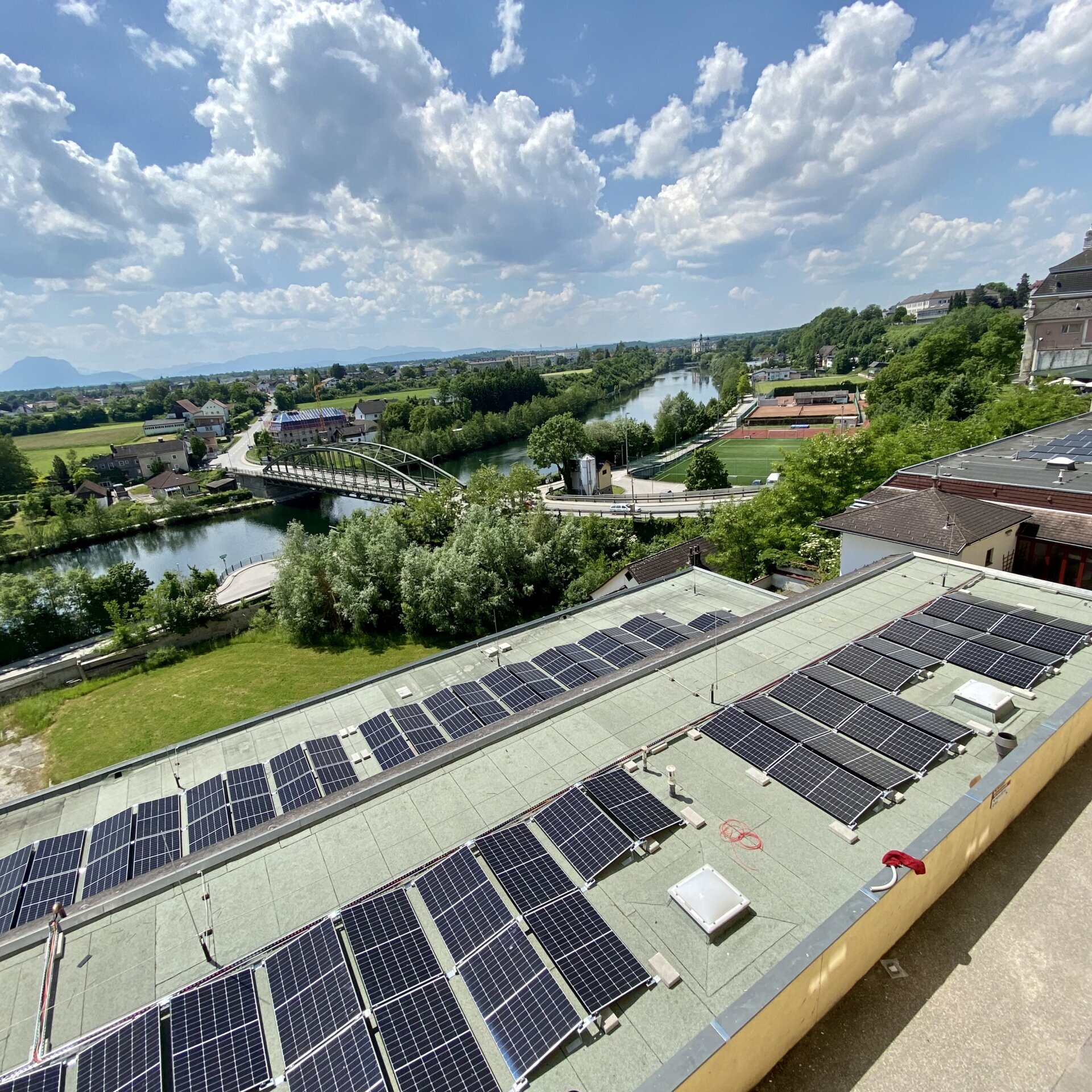 26-06-2023PV Anlage (14) Reihen von Solarmodulen auf einem flachen Dach überblicken einen Fluss, eine Brücke und die umliegenden Grünflächen unter einem teilweise bewölkten Himmel.