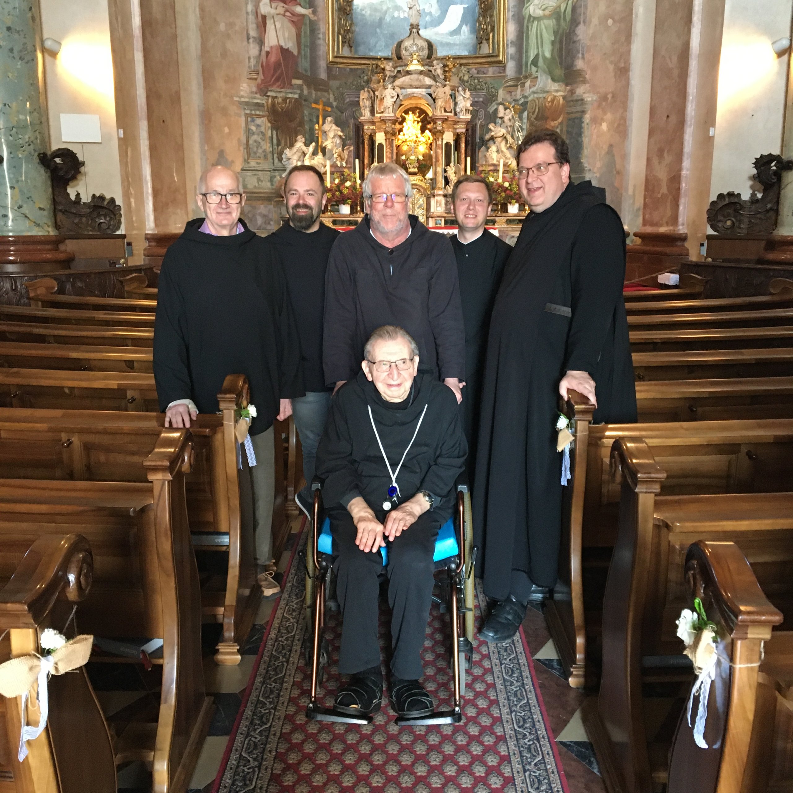 Sechs Männer, von denen einer im Rollstuhl sitzt, posieren für ein Gruppenfoto in einer Kirche mit verzierten Altären und Holzbänken.