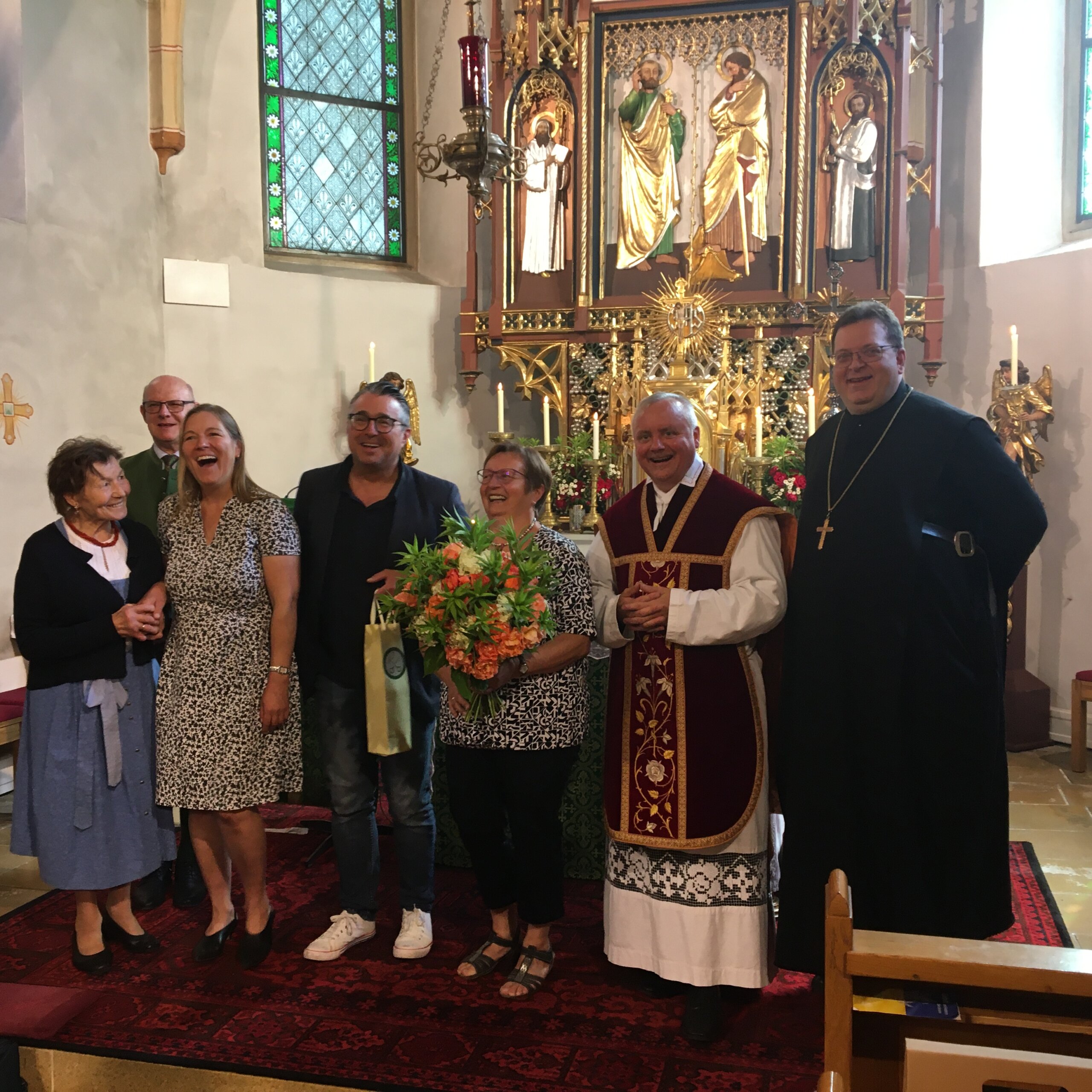 Eine Gruppe von sieben Personen, darunter Geistliche und Laien, steht in einer Kirche vor einem verzierten Altar zusammen; eine Person hält einen Blumenstrauß.