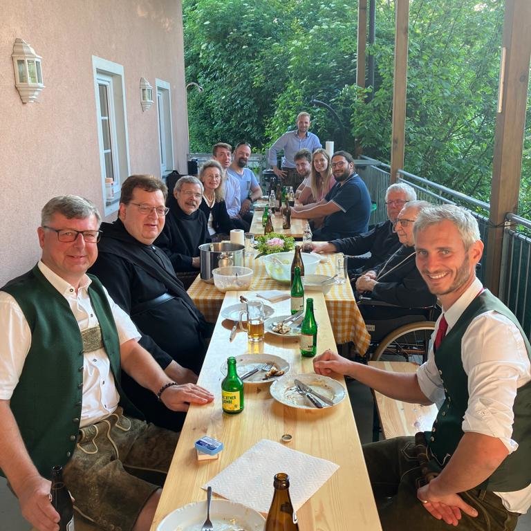 PHOTO-2023-06-12-20-26-19 Eine Gruppe von Menschen in traditioneller Kleidung sitzt um einen langen Holztisch auf einer überdachten Veranda und teilt eine Mahlzeit mit Getränken und leeren Tellern vor ihnen. Im Hintergrund sind Bäume und Sonnenlicht zu sehen.